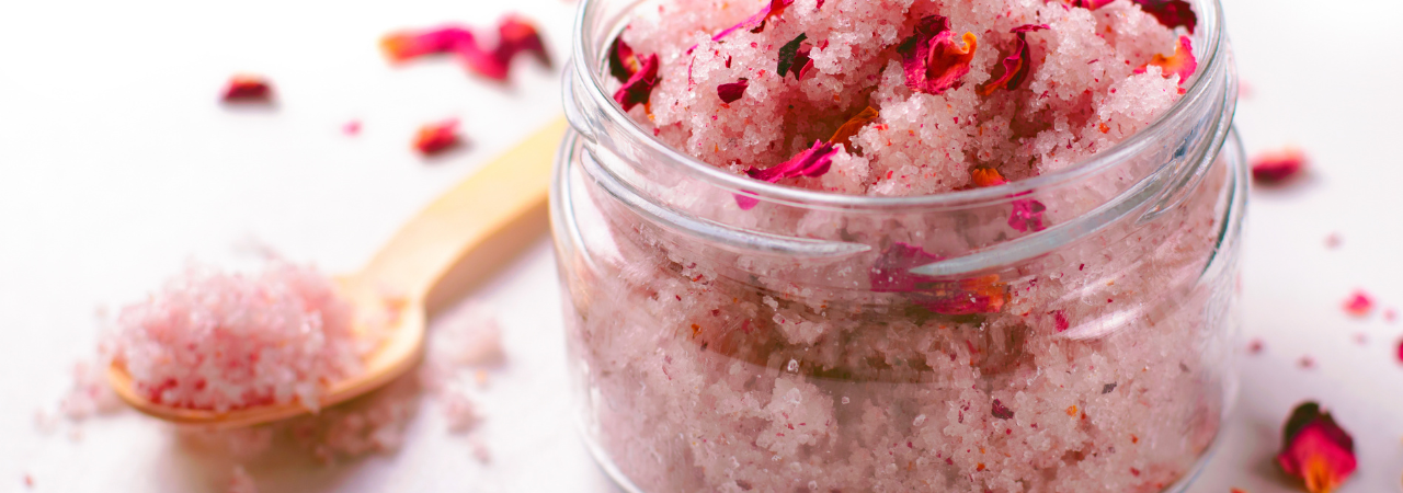 Close up of jar and spoon containing pink bath salts, with bright pink flower petals scattered